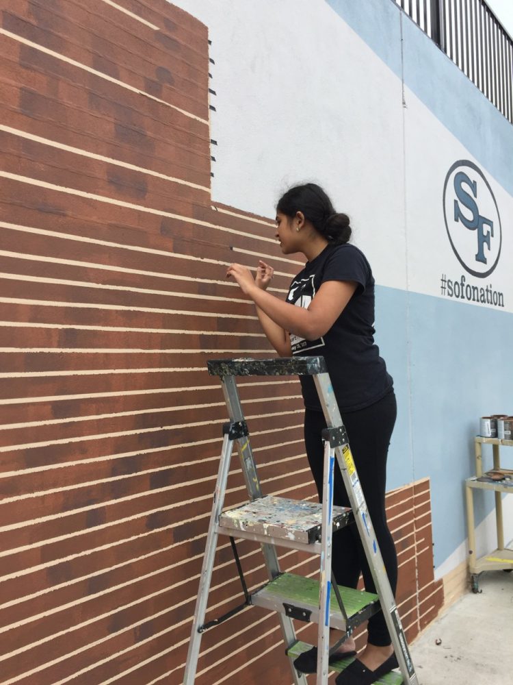 Here, Yanzhi Zhang paints the brick wall found right on the outside of the mural. Scott McIntyre told the Bird Feed Staff that the goal was to pull the mural back into the rest of the buildings, which are brick. Photo used with permission from Scott McIntyre.