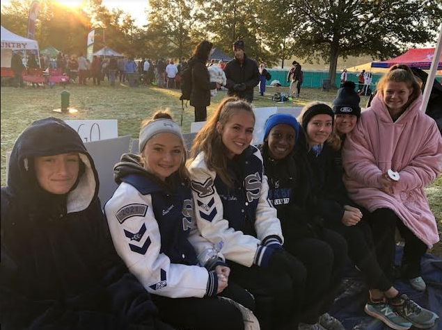 Bella Rhone, Emma OConnor, Carmel Yonas, Sophia Baker, Sydney Copeland, Madelynne Cadeau, and Caroline Patterson sit together covered up. On the morning of state on a Saturday, varsity girls warm themselves up to be ready for the race. They are determined to do well and are going to rely on all the training theyve had to prepare for state. 