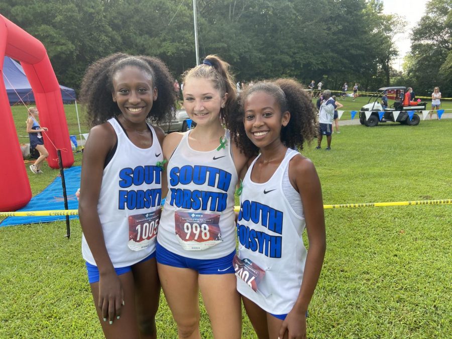 3 varsity girls. On August 22, Carmel Yonas (Sophomore), Emma OConnor (Senior), and Isabel Yonas (Freshman) finish the race smiling as they take a group picture. The girls wore a green ribbon in memory of Trevor Lockhurst who went to South, and was on the Cross-country team. 