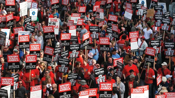 A crowd, filled with both creators and viewers, hold up signs of protest. Many display "Solidarity With Writers," or "Writer's Guild of America: On Strike."  